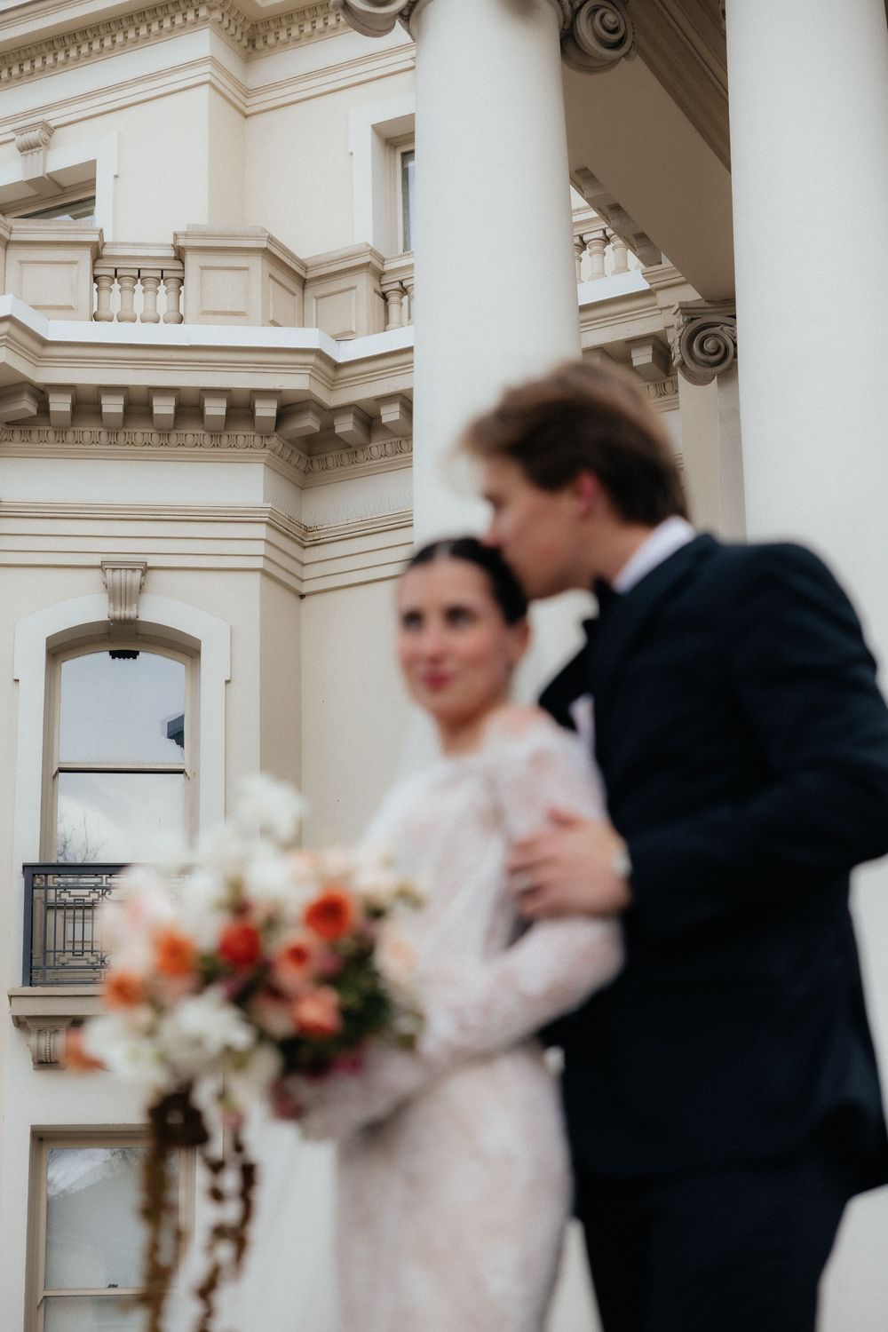 Groom kisses bride near grand white columns of elegant venue, bride holding colorful peach bouquet.