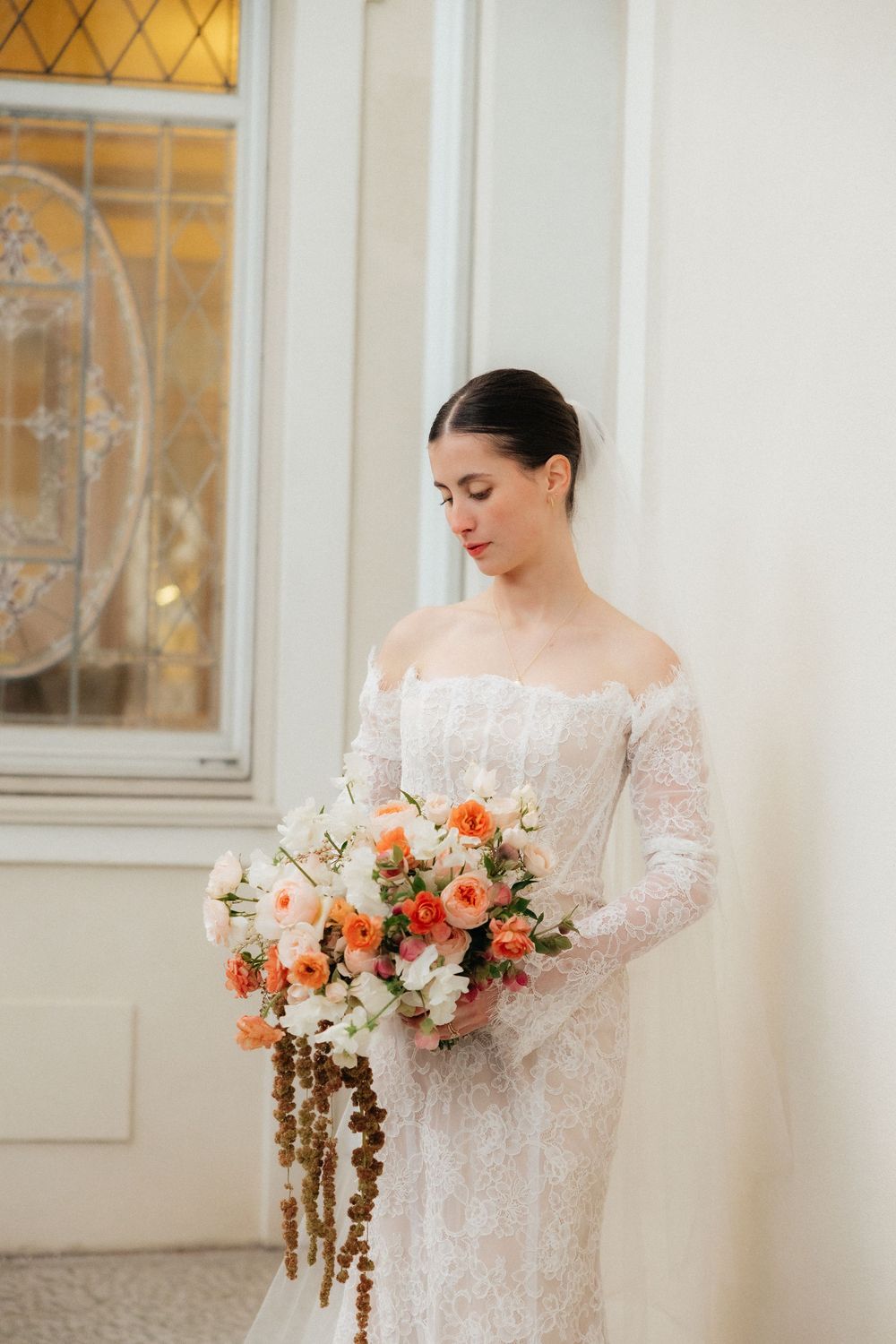Bride in off-shoulder lace gown gazes down at cascading peach and white bouquet by stained glass window.
