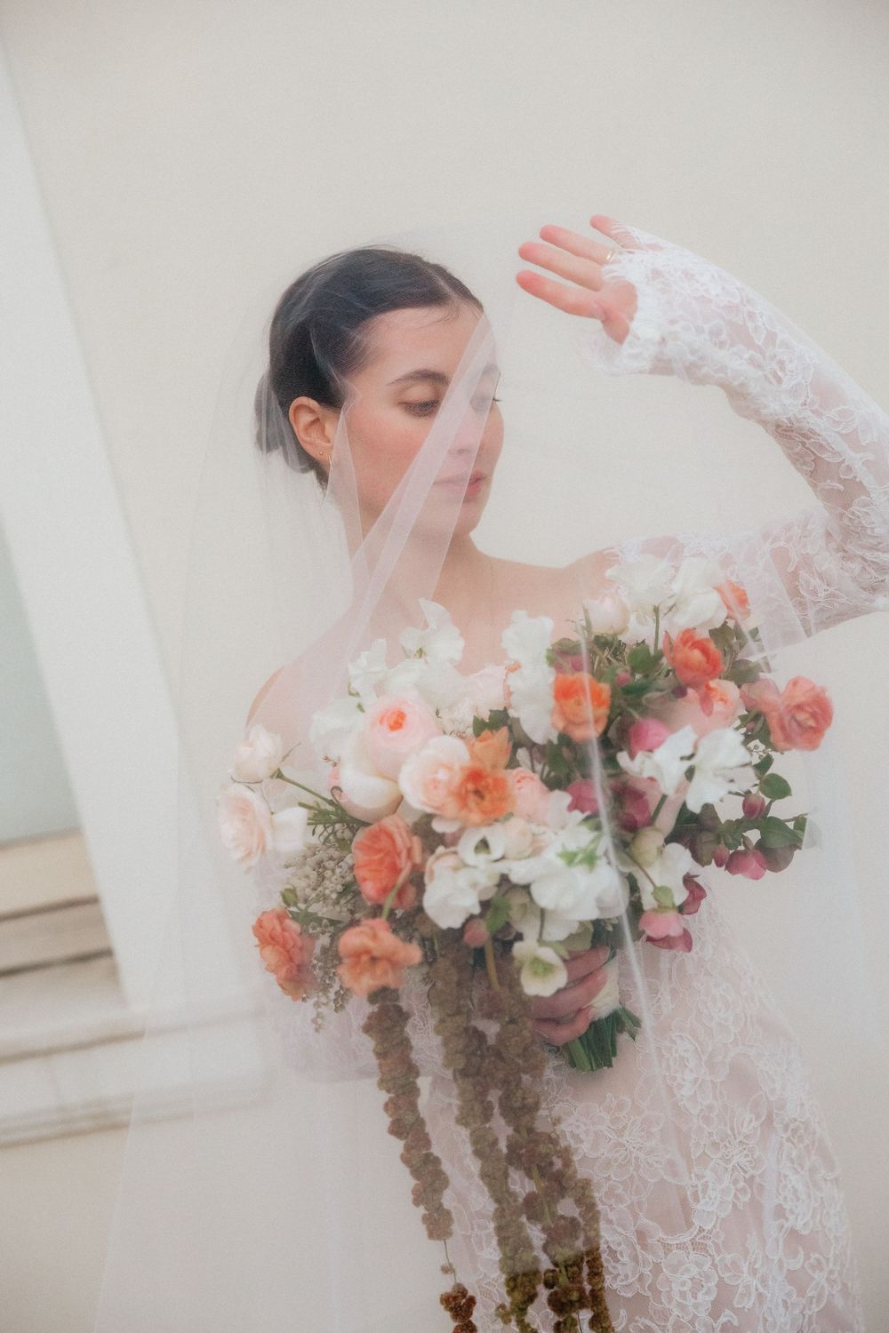 Bride in lace gown holds coral and white bouquet, draped in sheer veil, posing elegantly against a soft neutral background.