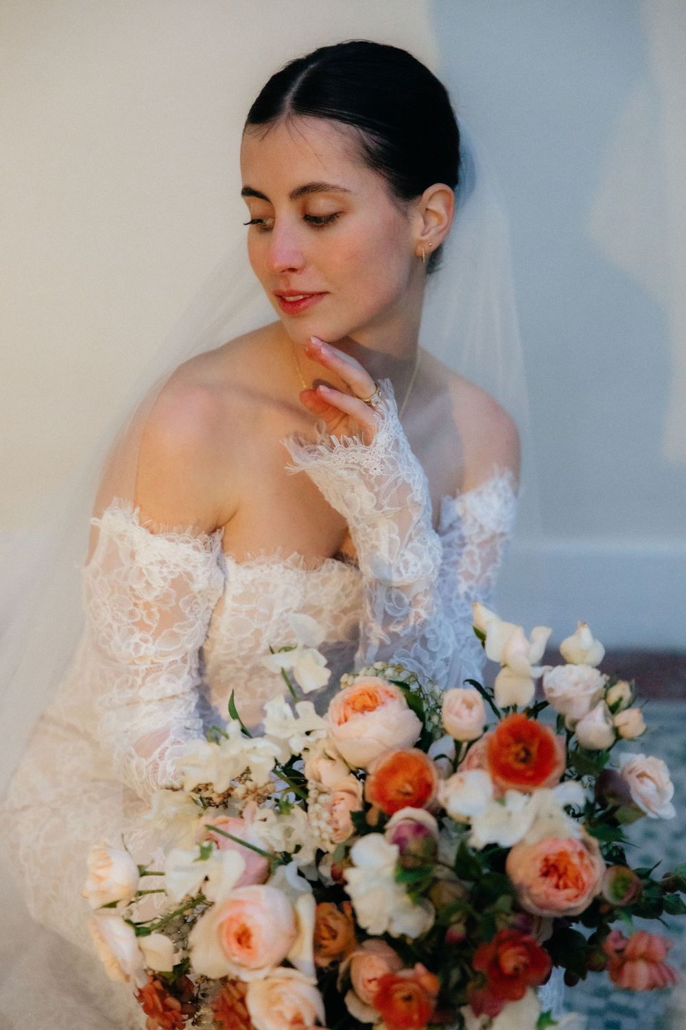 Bride in off-shoulder lace gown holds colorful bouquet of peach and white roses, eyes closed, serene expression.
