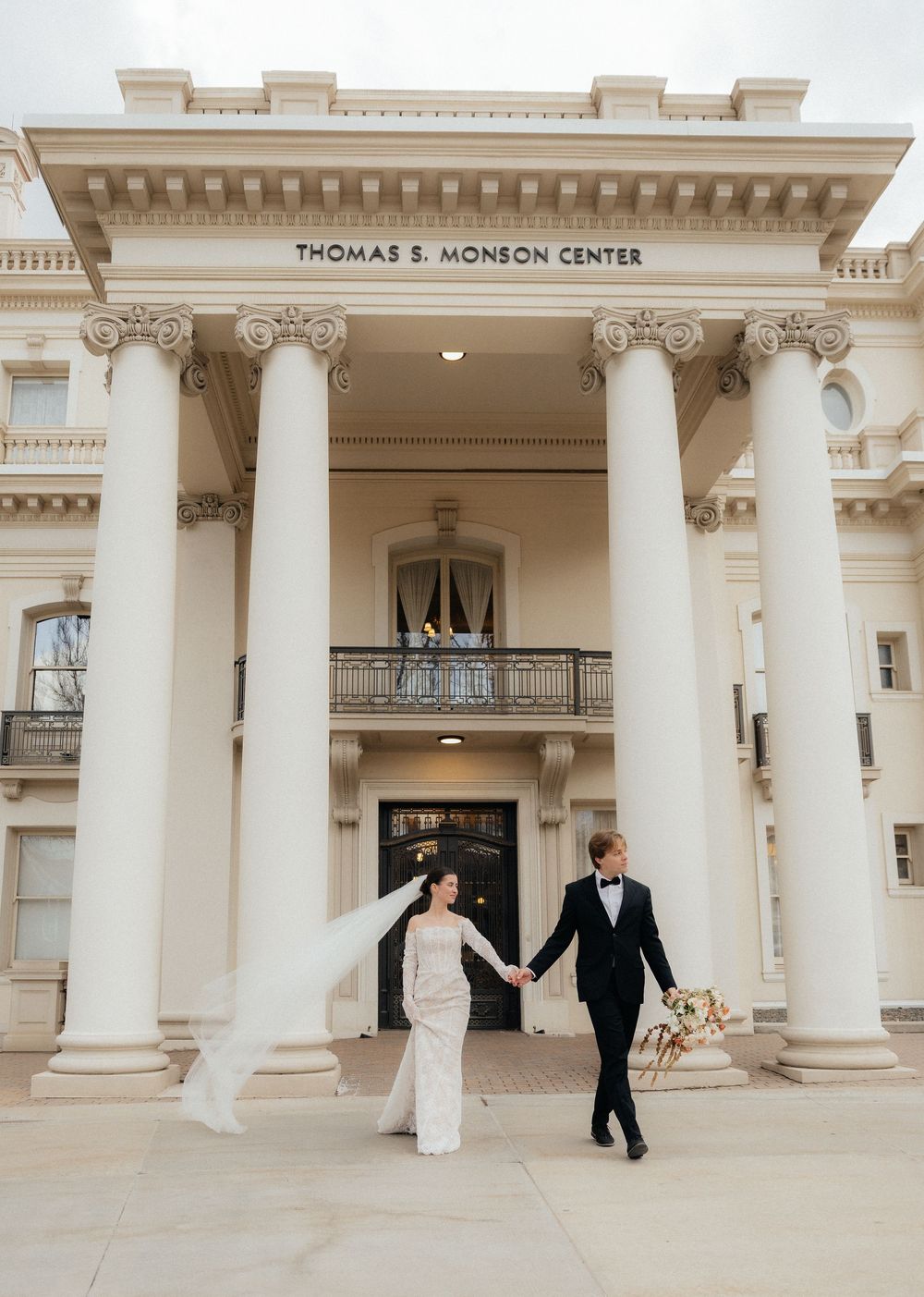 Wedding couple holds hands walking before Thomas S. Monson Center with grand white columns and neoclassical architecture.