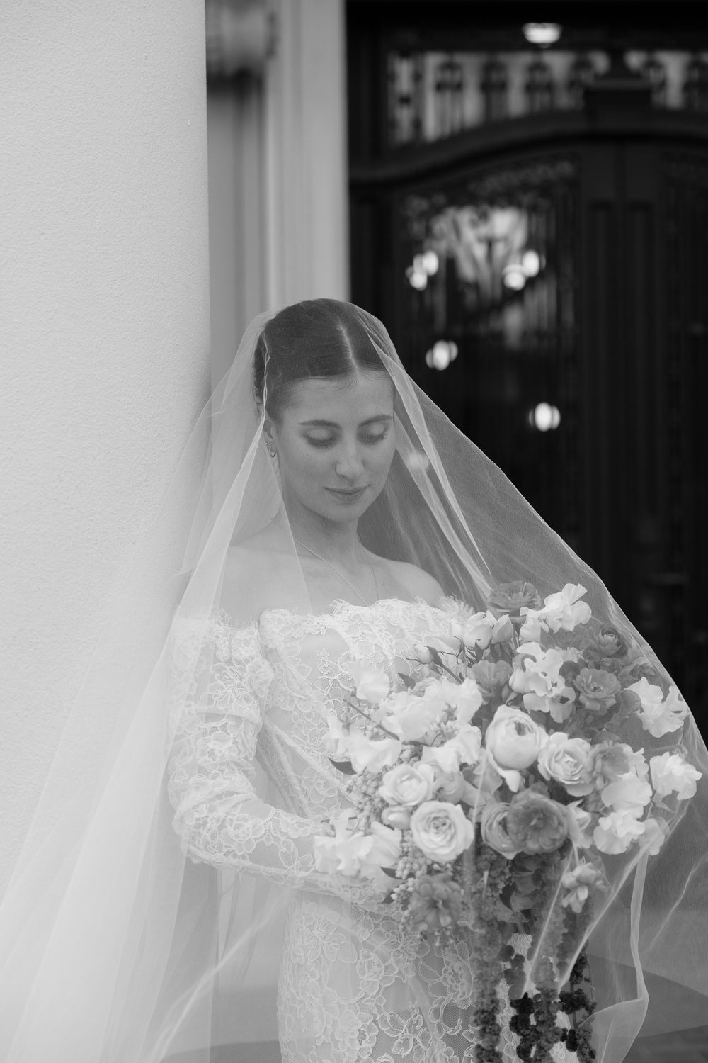 Black and white portrait of bride in lace gown and veil holding floral bouquet by ornate wooden door.