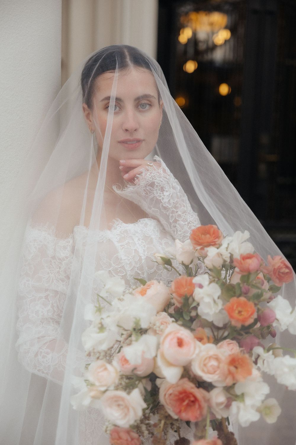 Bride in lace off-shoulder gown holds peach floral bouquet under flowing veil, posing elegantly indoors.