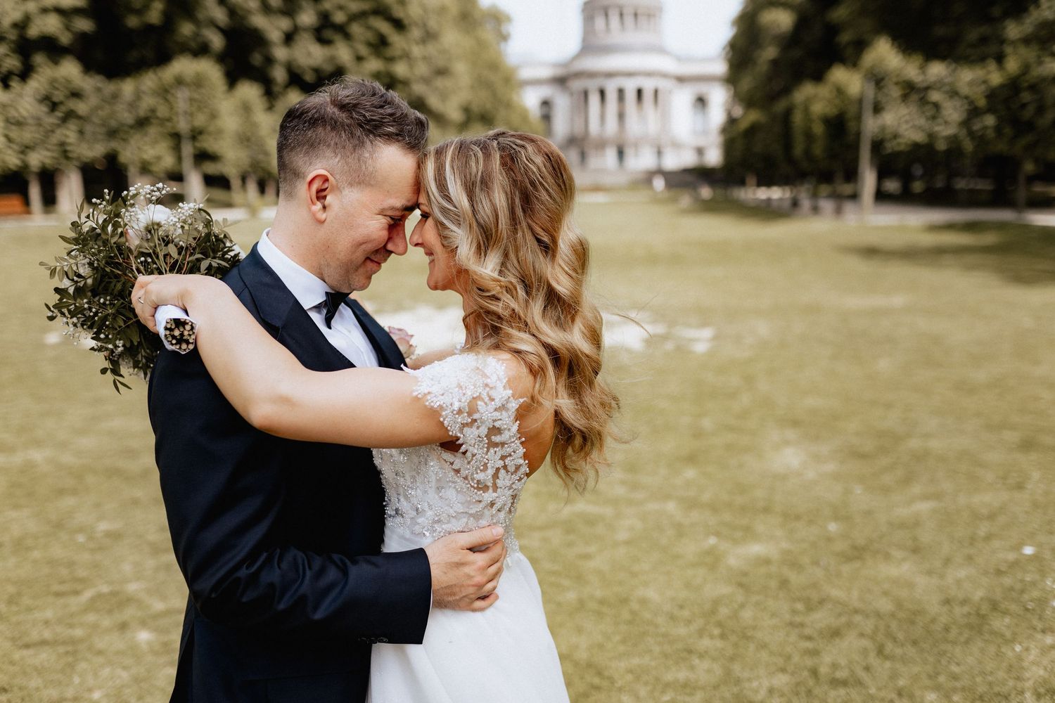 A bride and groom embrace outdoors on a green lawn, with a domed building in the background on their wedding day.