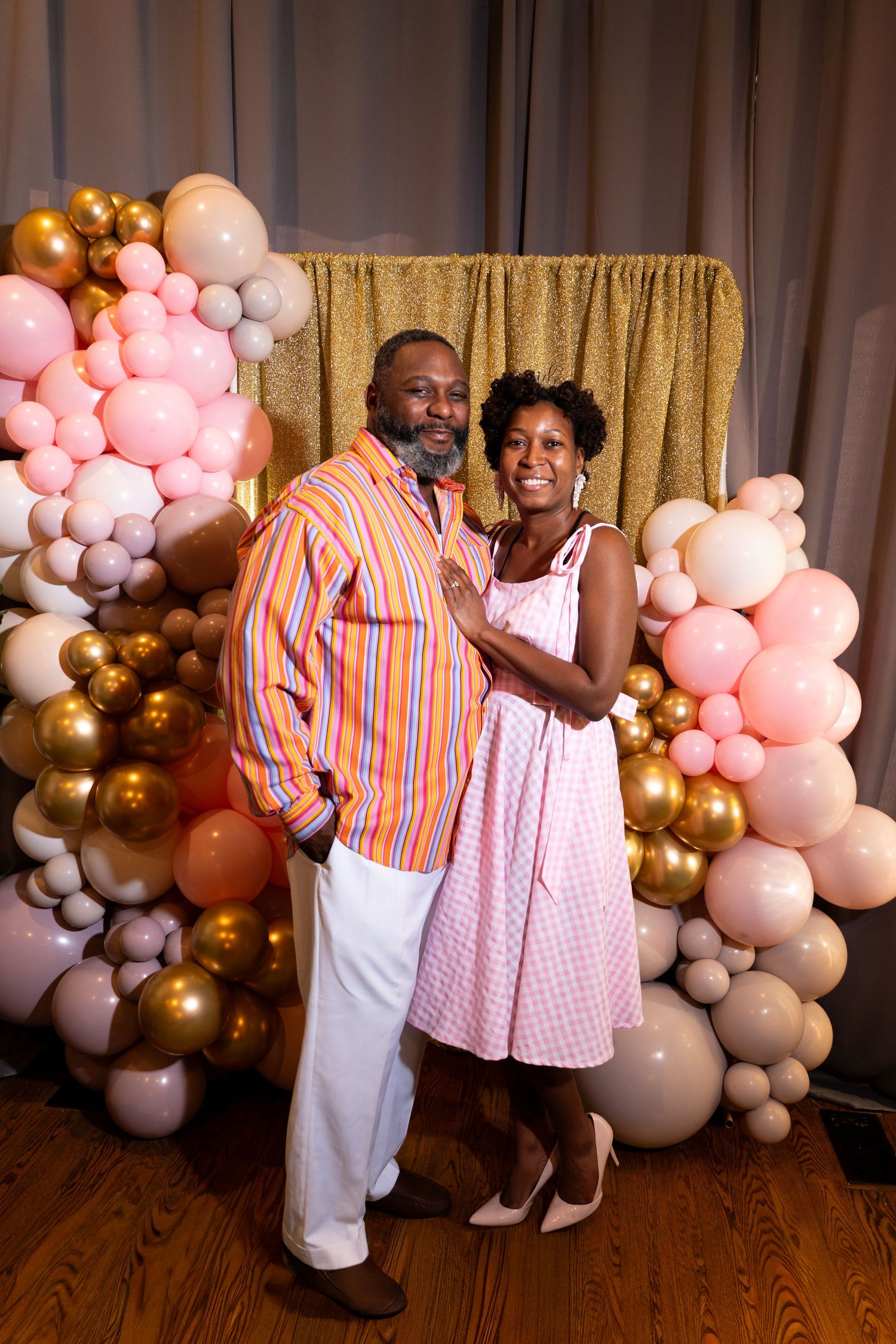 Couple poses together in front of pink and gold balloon backdrop at a celebration event.