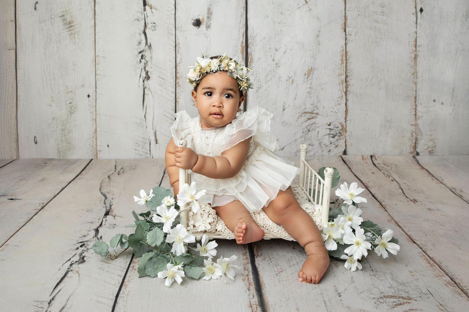 Baby girl in white lace dress and floral crown sits beside a miniature white bed with flowers on rustic wood floor.