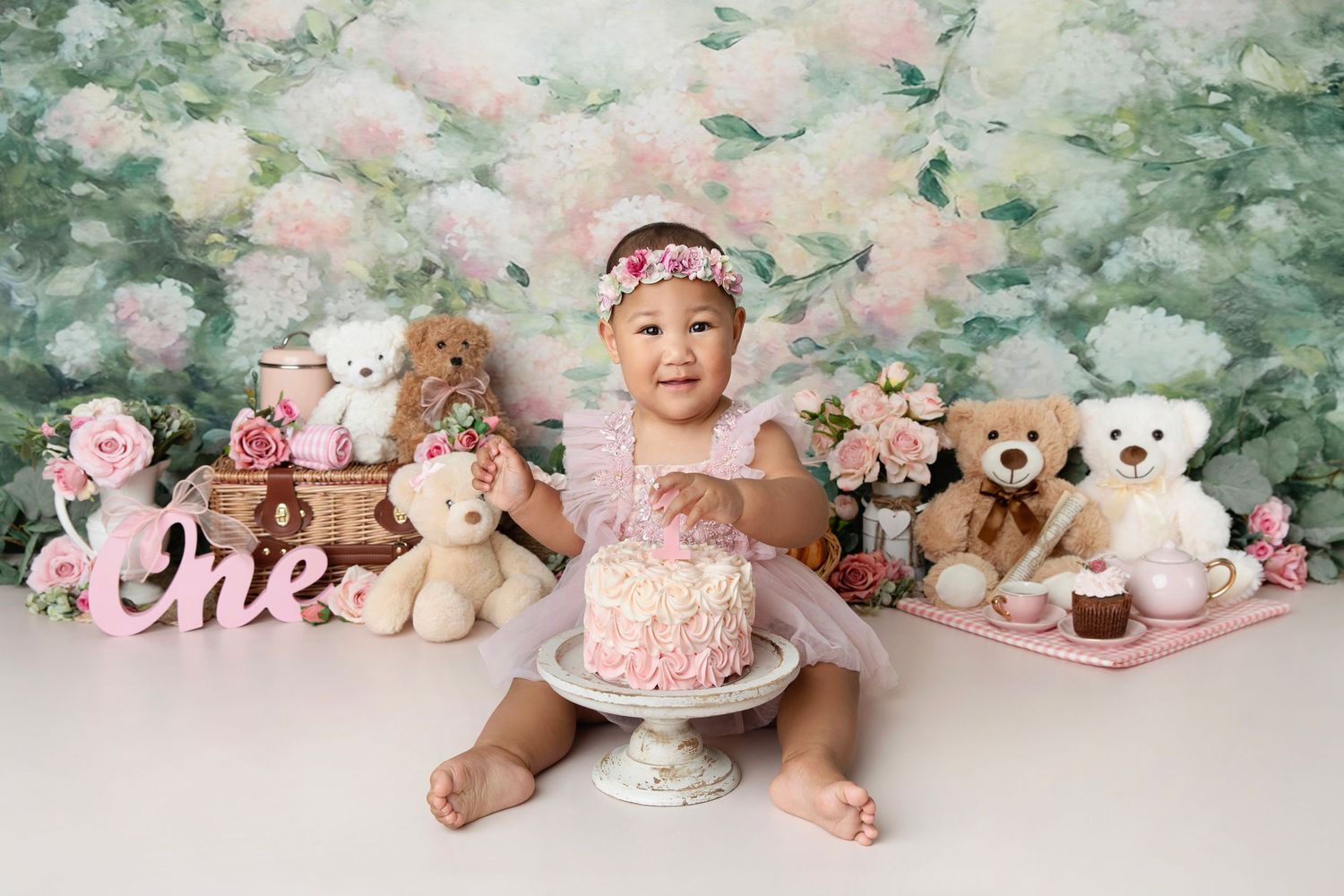 Baby girl in pink tutu celebrates first birthday with cake, teddy bears, and floral backdrop.