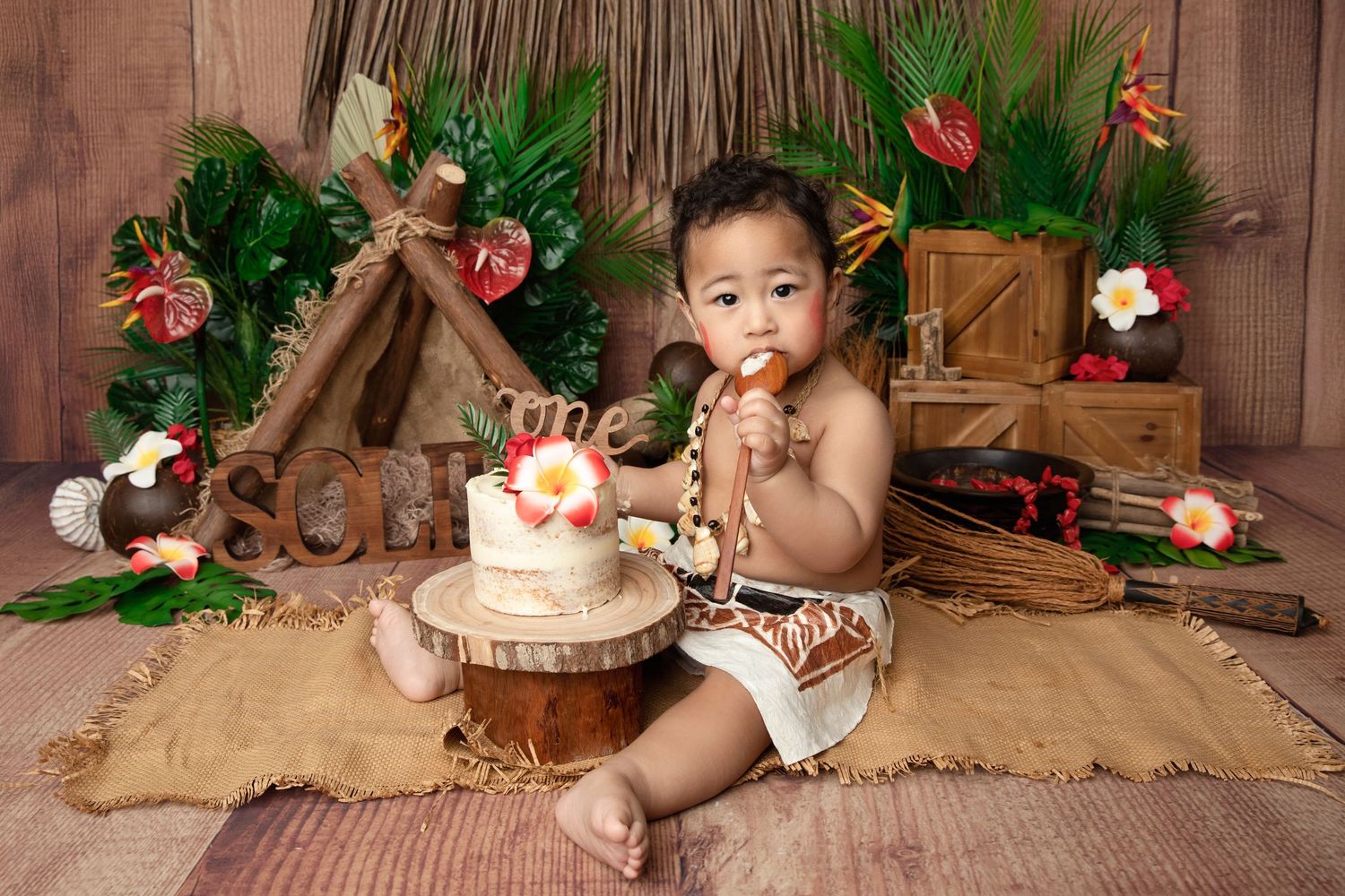 Baby boy in tribal outfit sits beside a small cake on a wood stump, surrounded by tropical decor and greenery.