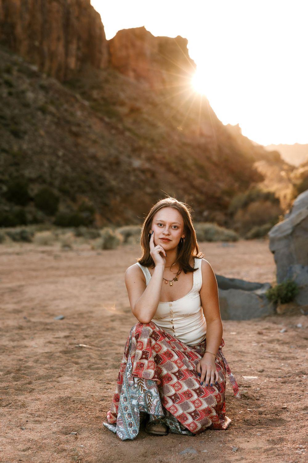 Young woman in bohemian skirt sitting in desert landscape with golden sunset behind rocky mountains.