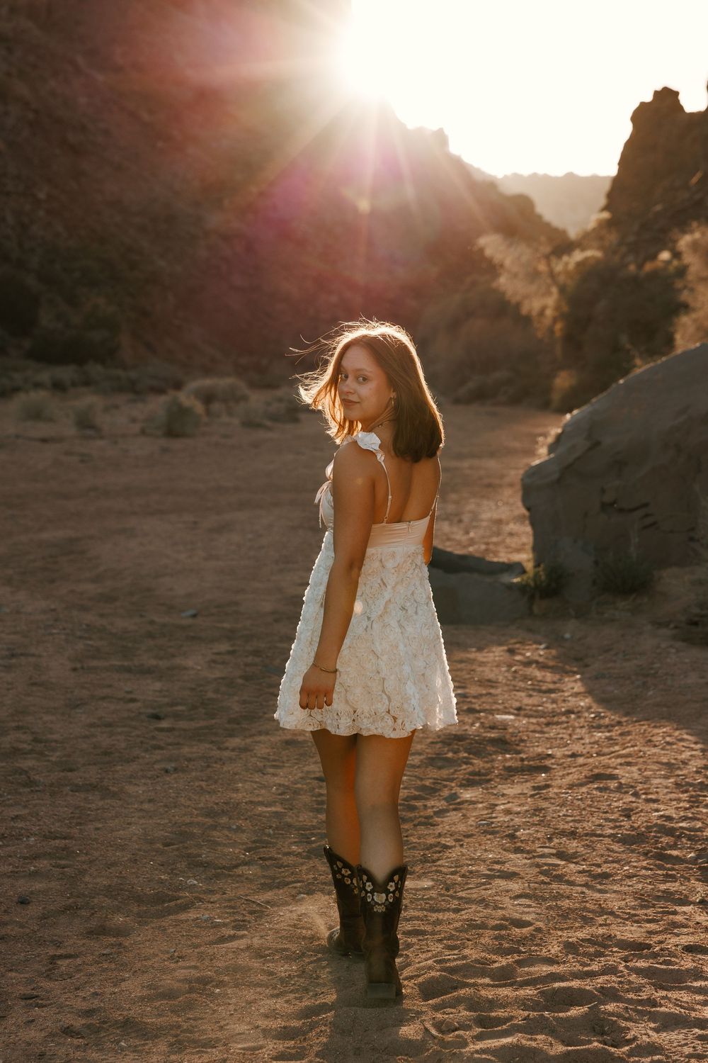 Woman in white lace dress and cowboy boots stands in desert canyon at sunset.