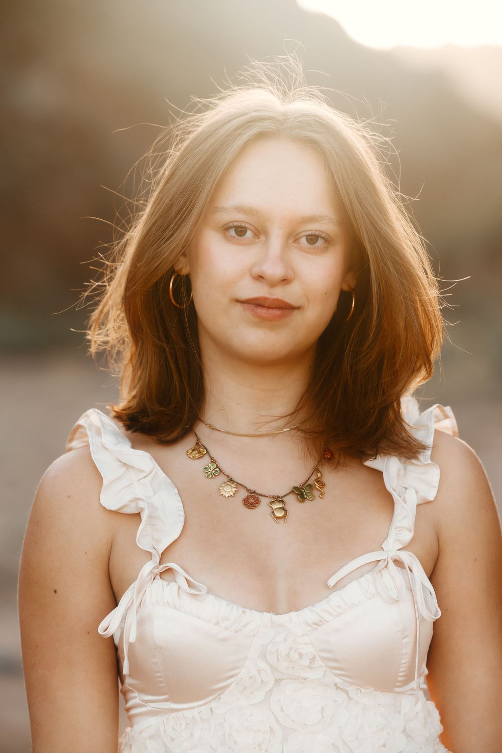 Portrait of young woman with auburn hair wearing white ruffled dress and gold necklace at sunset.