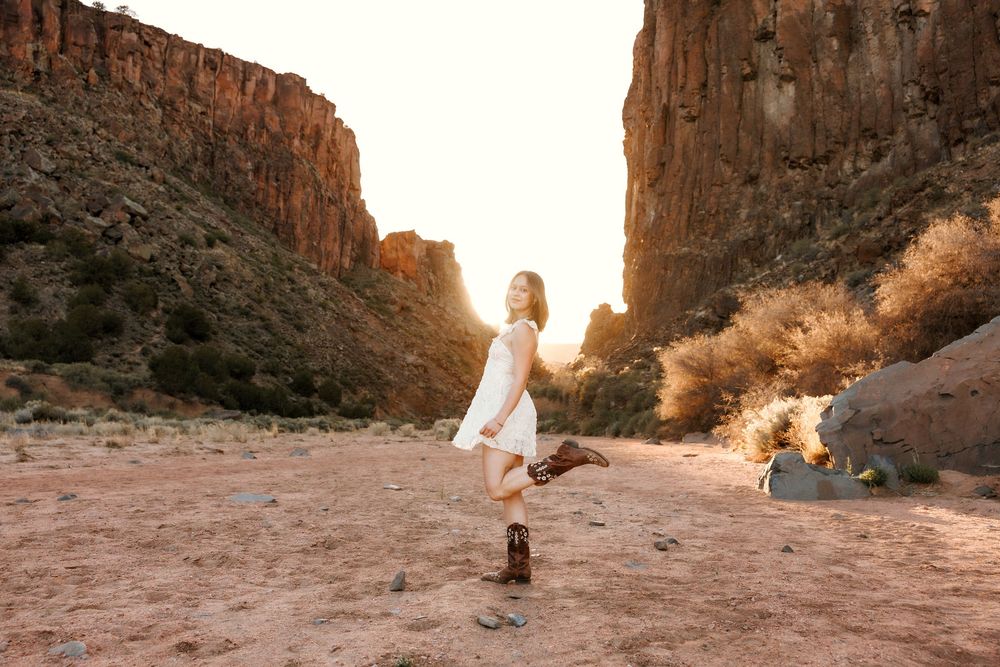 Woman in white dress and cowboy boots poses gracefully in a stunning red rock canyon at golden hour.