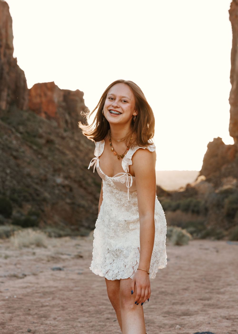 Smiling young woman in white lace mini dress posing in desert canyon at sunset.