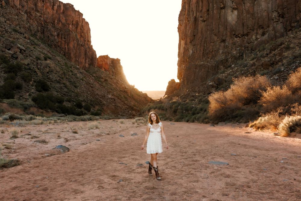 Young woman in white dress and boots walking through scenic desert canyon at sunset.