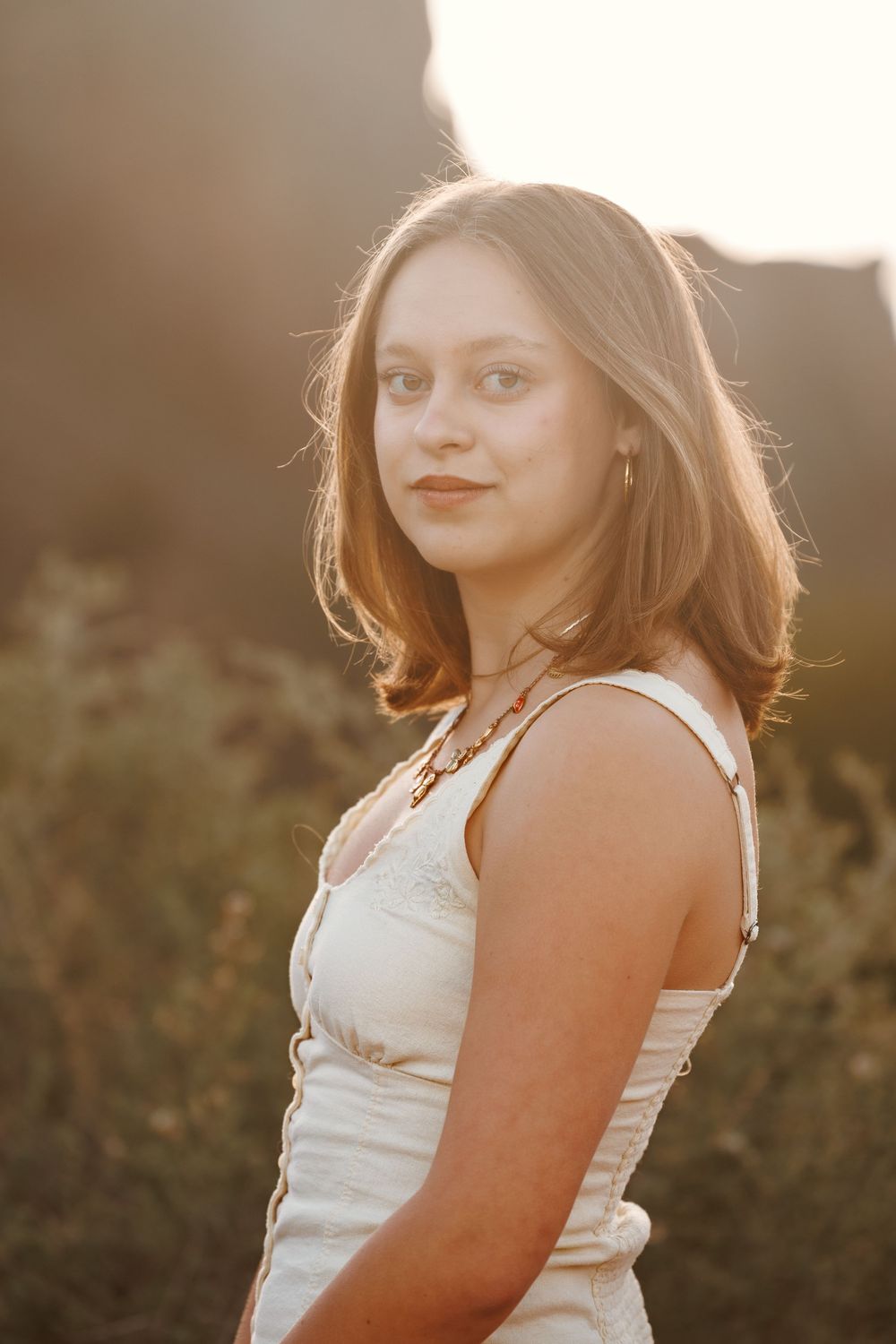 Young woman with auburn hair in white dress poses outdoors at golden hour sunset.