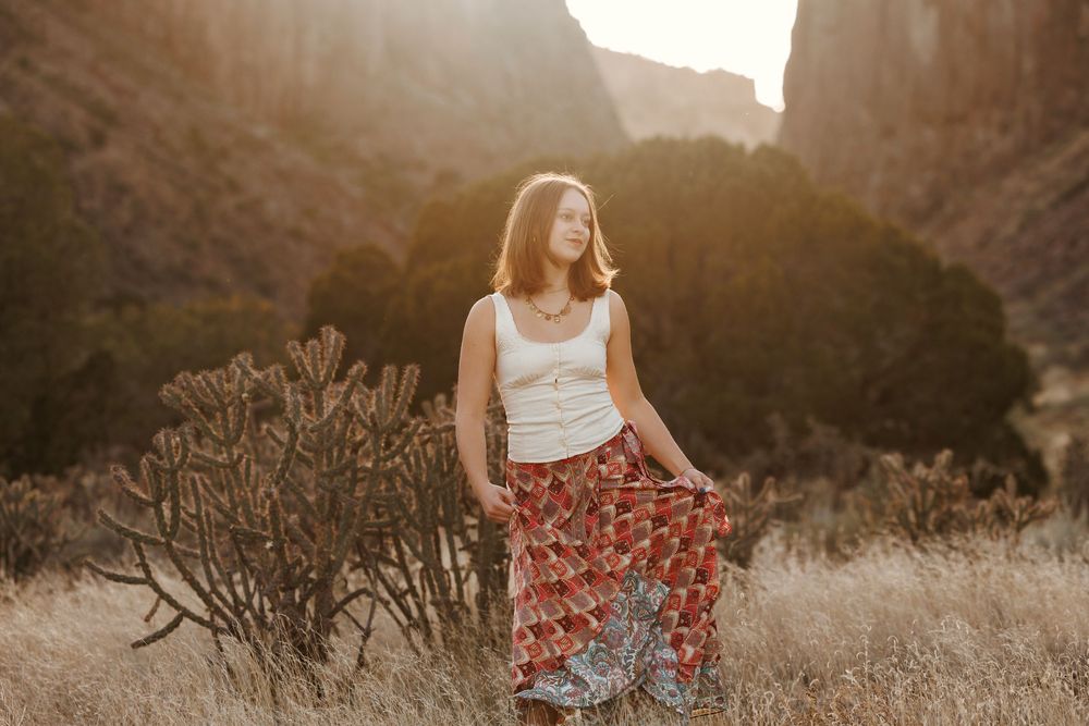 Young woman in floral maxi skirt and white top poses in golden desert landscape with cacti and canyon walls.
