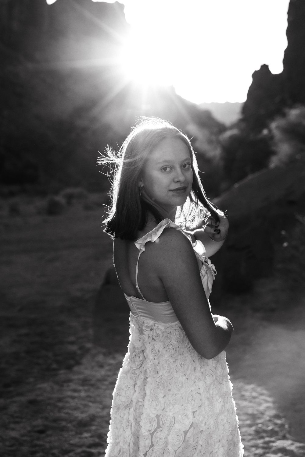 Dramatic black and white portrait of young woman in white dress with sun flare behind red rock canyon formations.
