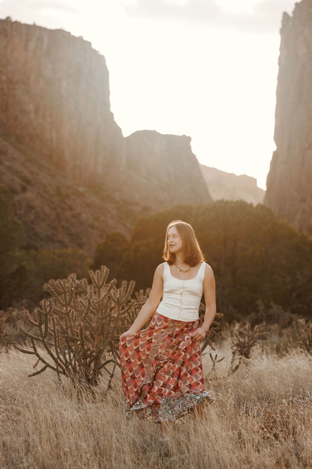 Young woman in white top and floral skirt standing in desert canyon at golden hour.