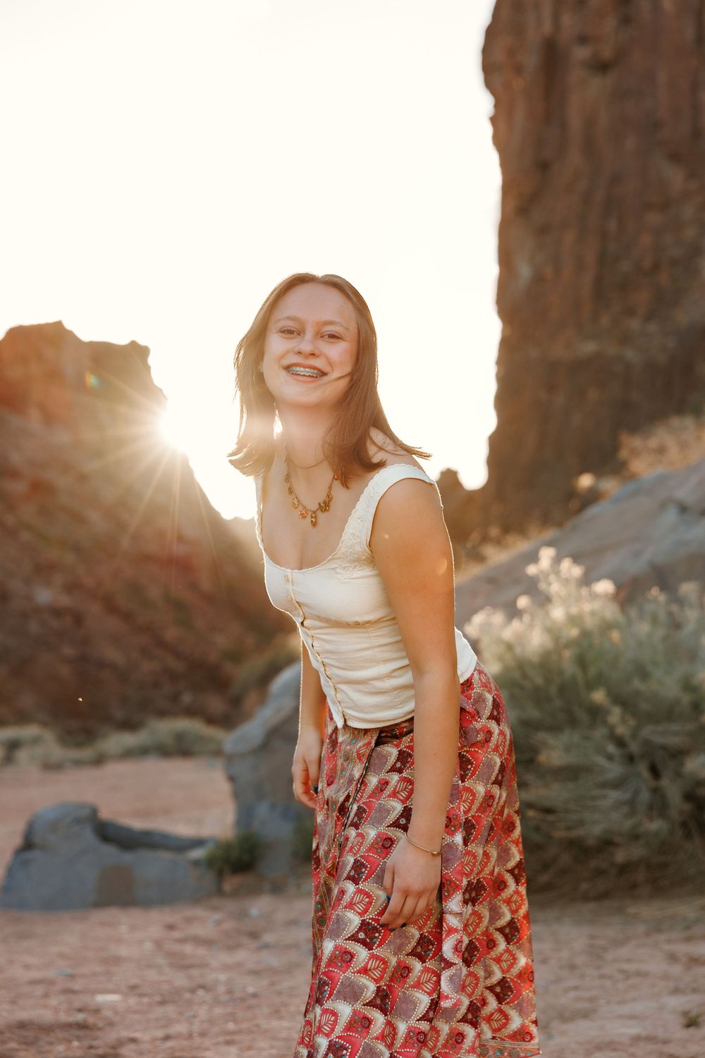 Young woman in white top and floral skirt smiling at golden hour near desert rock formations.