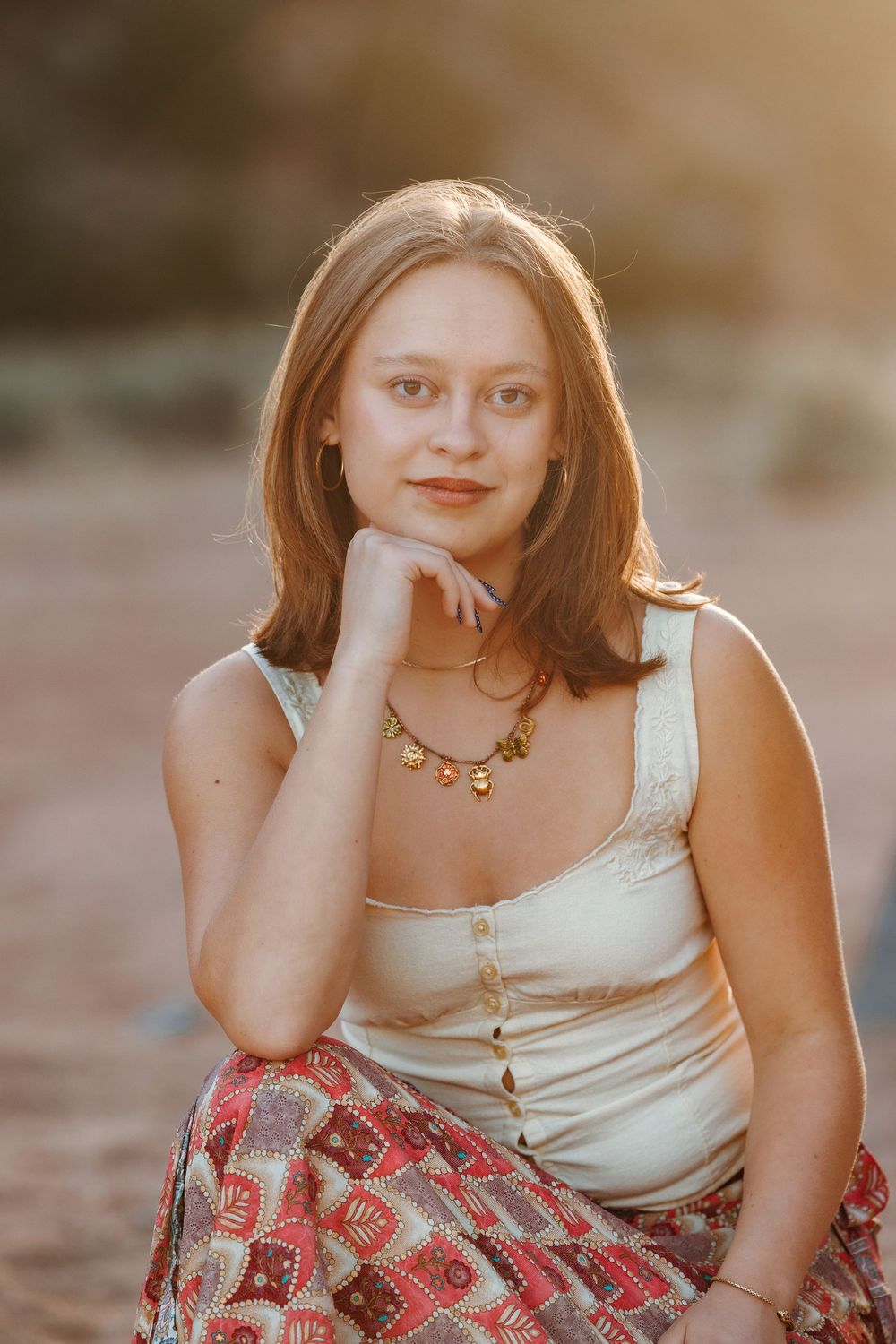 Young woman in white top and red patterned skirt poses with chin resting on hand during golden hour outdoor portrait.