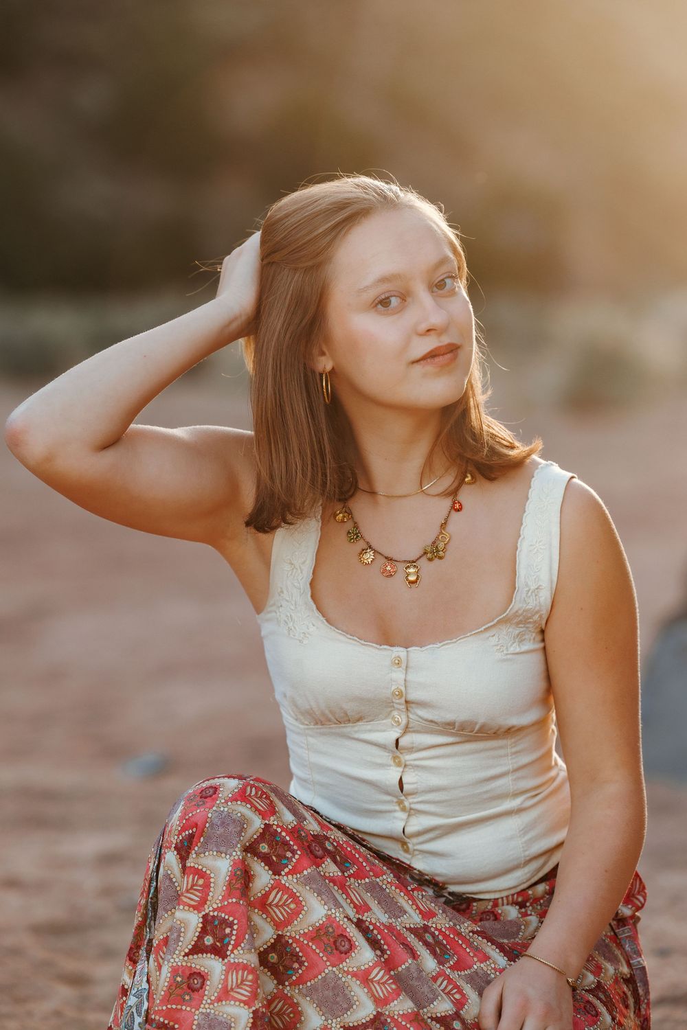 Young woman in boho white tank top and red skirt touches her hair during a warm sunset portrait session.