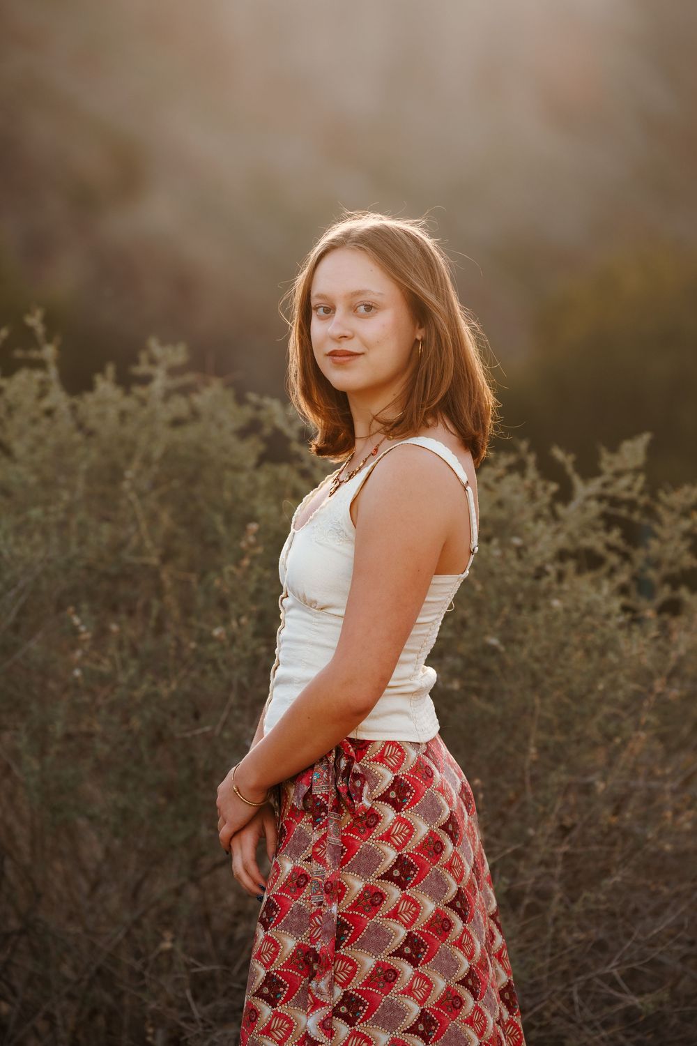 Young woman in white sleeveless top and red patterned skirt stands among desert shrubs in golden sunset light.