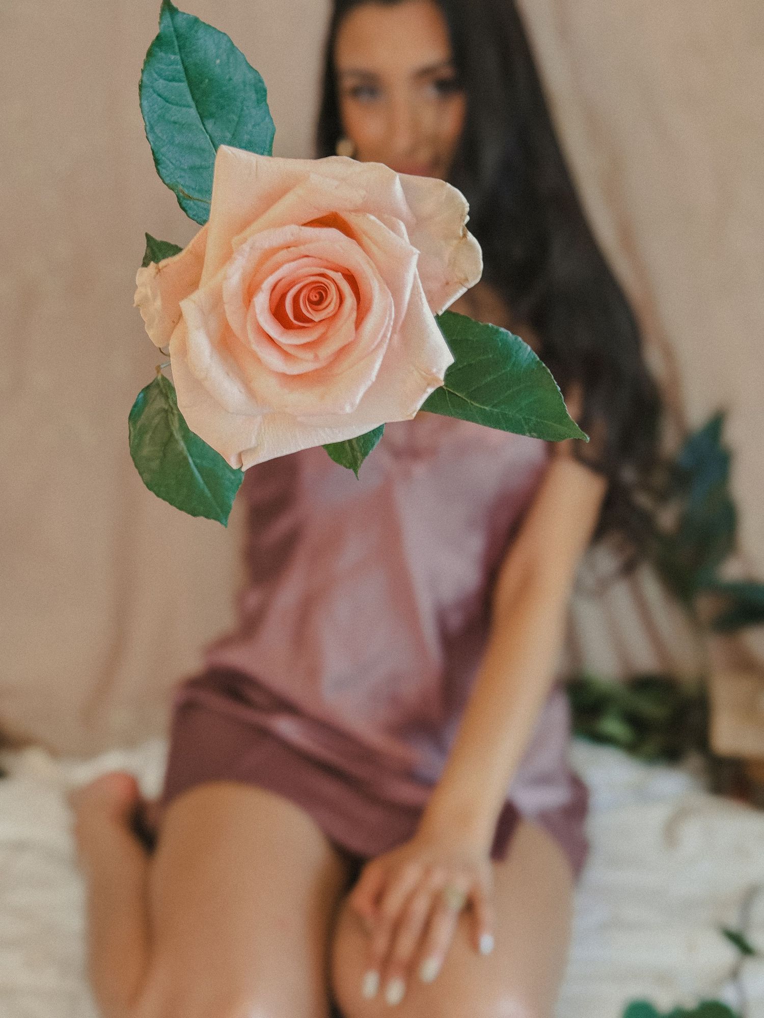 A peach rose held close to camera with blurred woman in mauve silk dress sitting on bed in background.