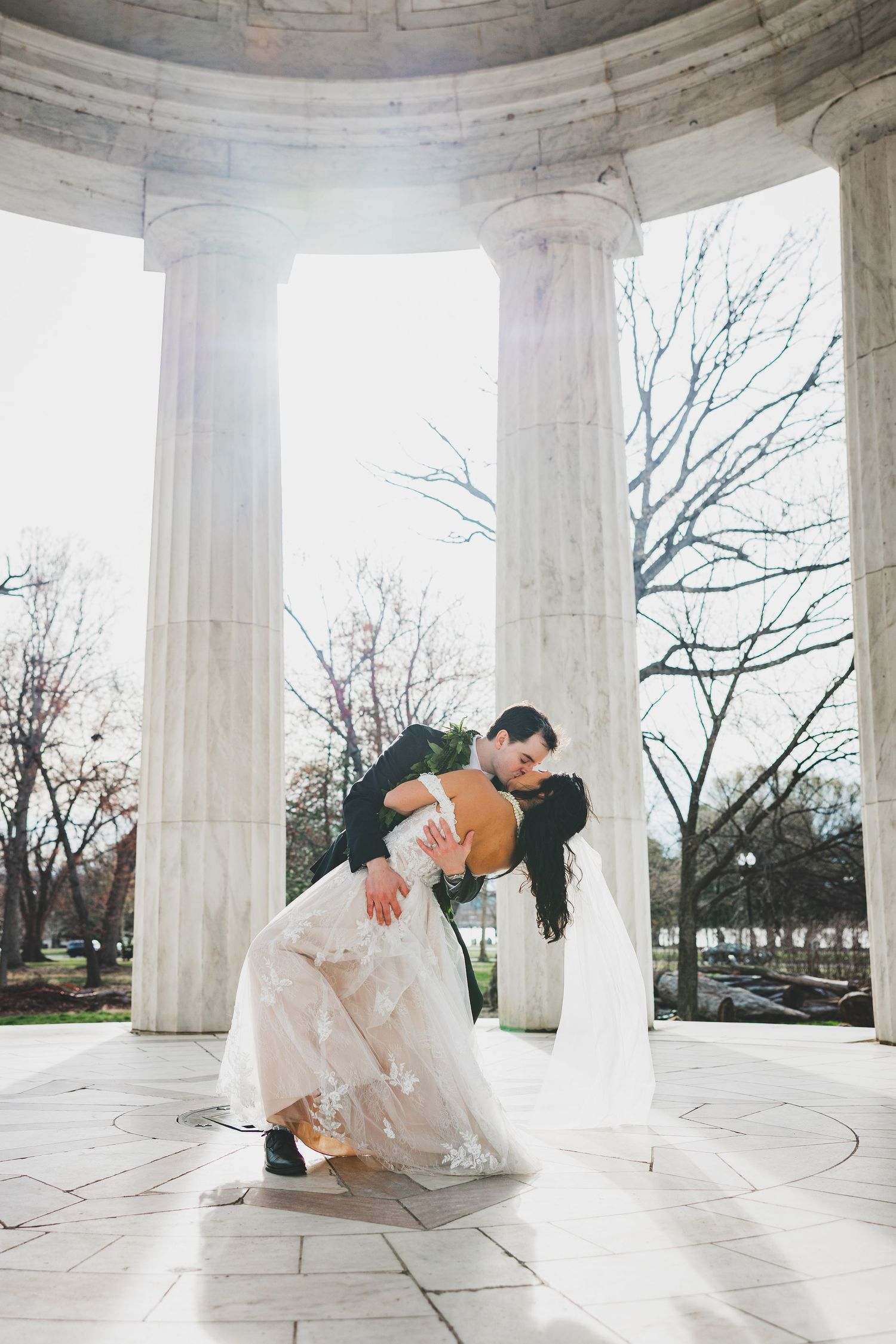Bride and groom share a romantic dip kiss under marble columns at a DC War Memorial wedding venue.