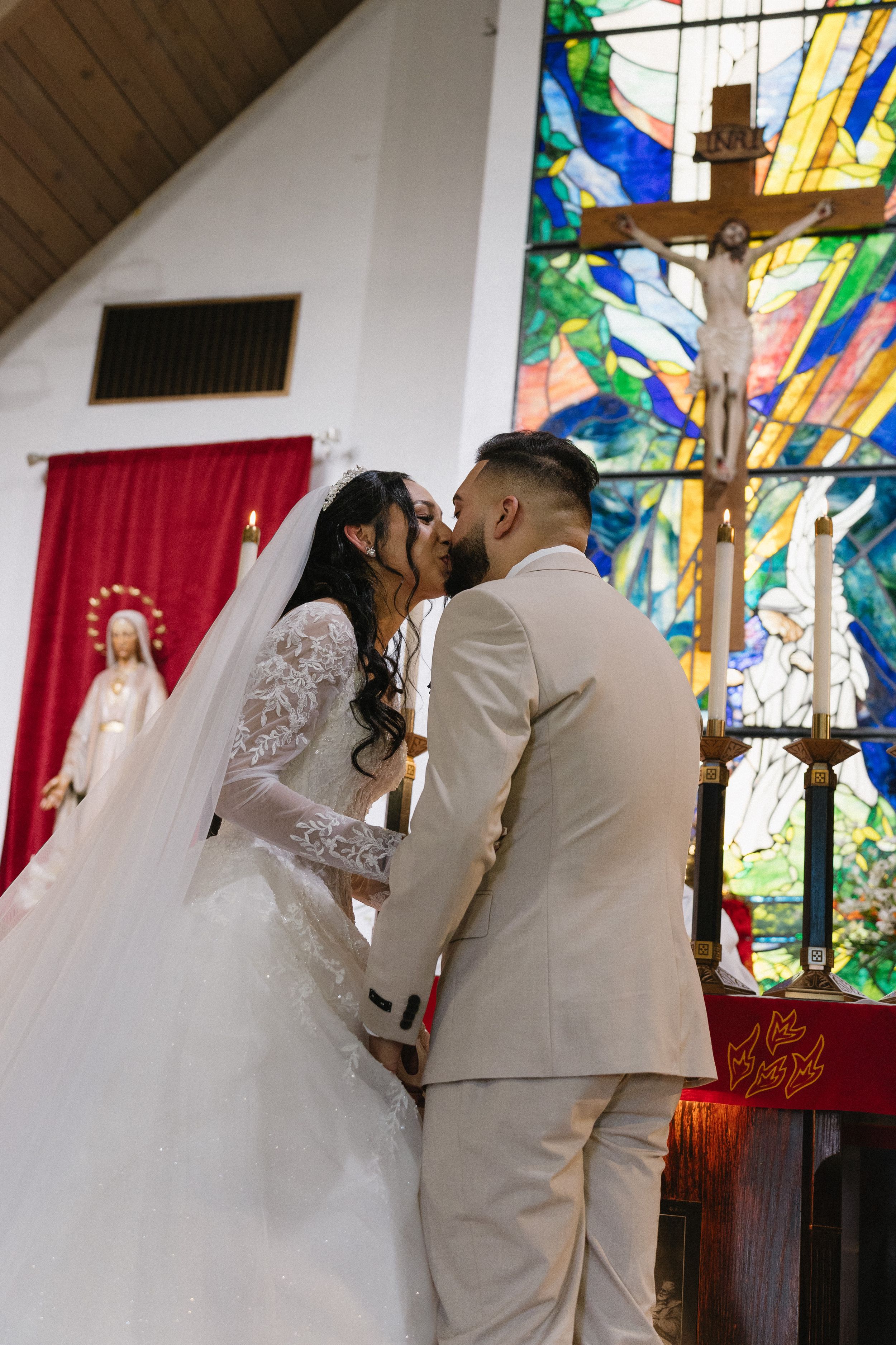 Bride and groom share first kiss at church altar with stained glass window and crucifix backdrop during wedding ceremony.