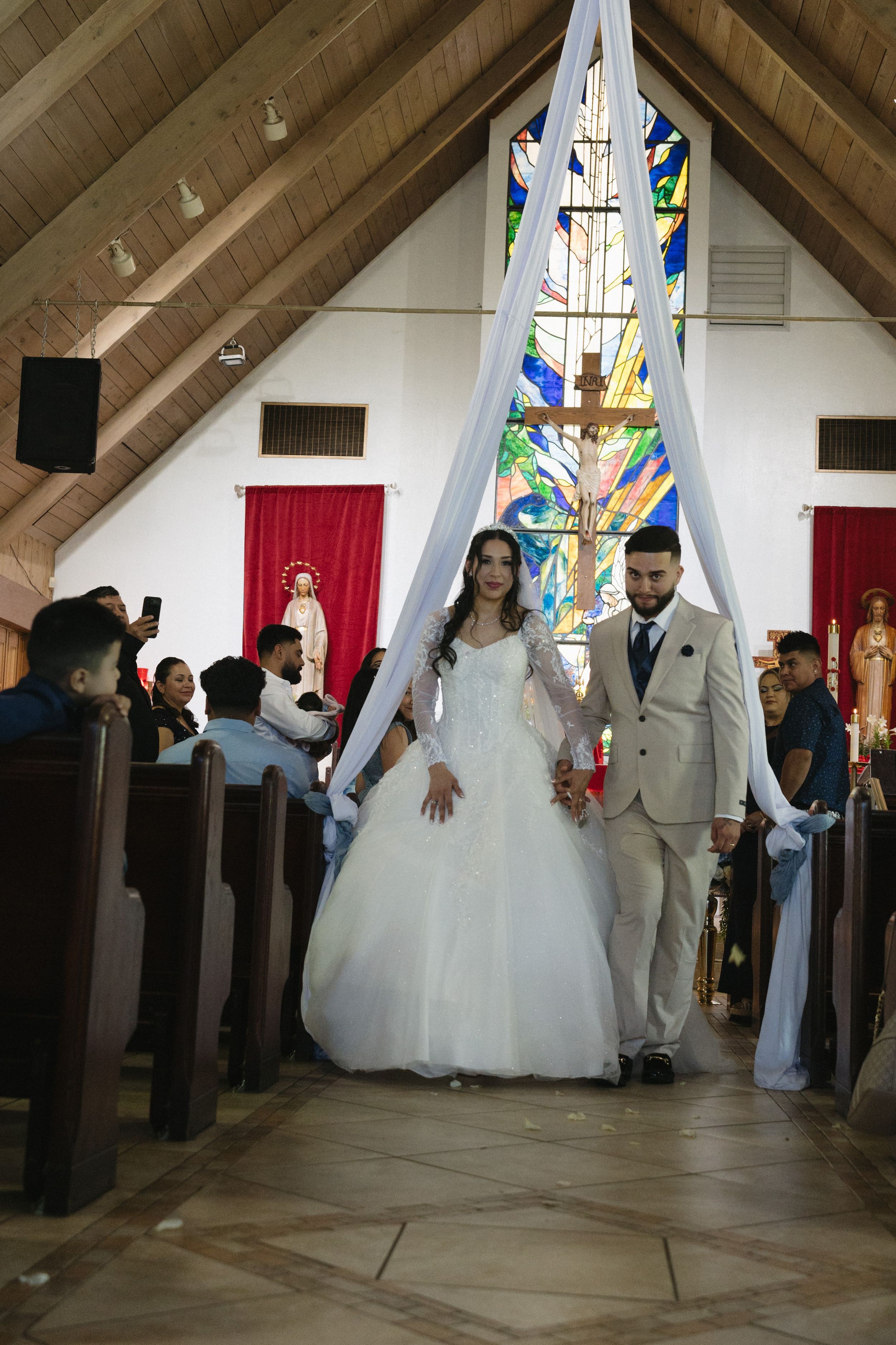Bride in white gown and groom in beige suit walk down church aisle after wedding ceremony with stained glass backdrop.