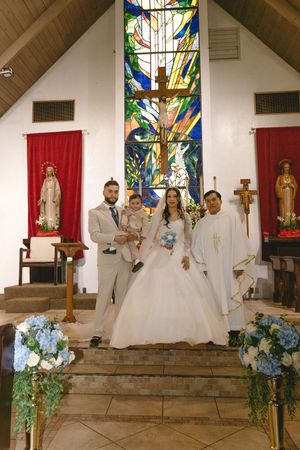 Wedding couple poses with priest and child at church altar featuring colorful stained glass window.