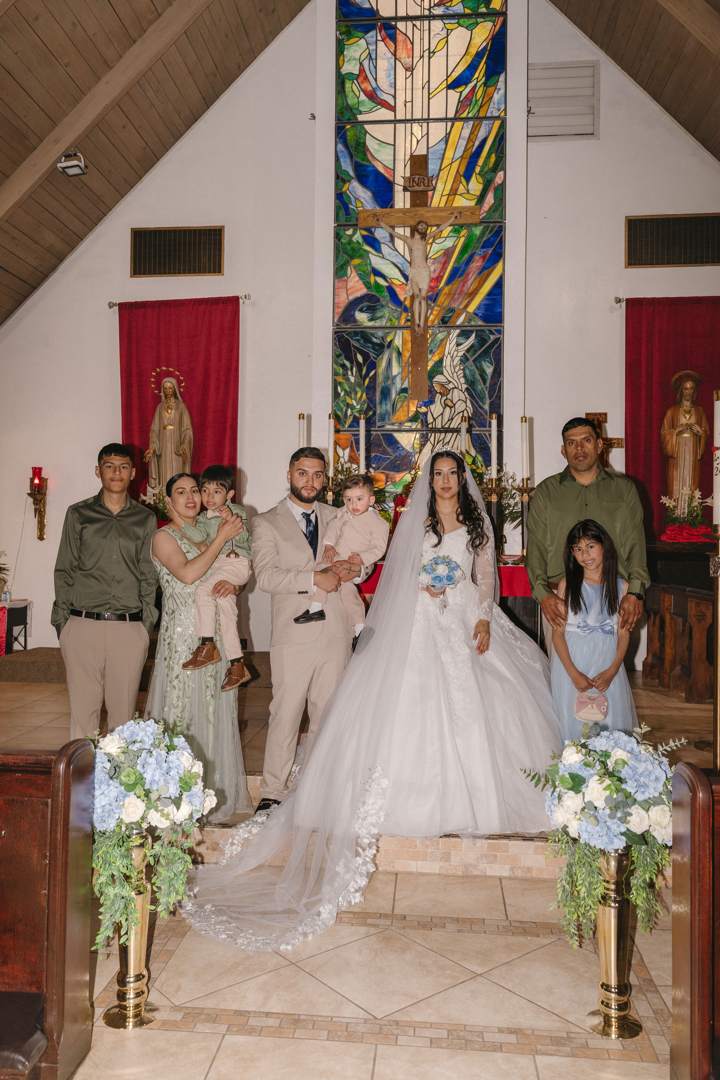 Wedding party poses at church altar with stained glass cross, bride in white gown holding bouquet, groom in tan suit.