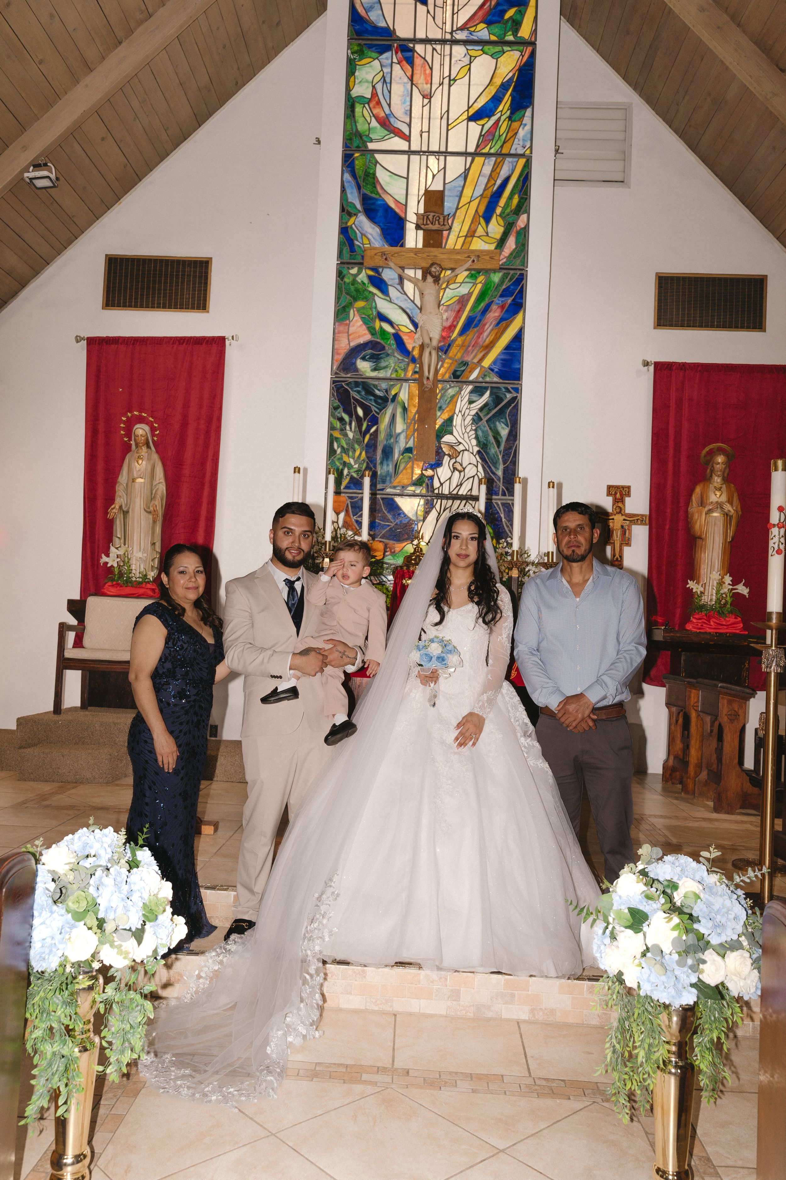 Bride and groom pose with family in a church altar, bride in white gown holding bouquet, stained glass window backdrop.