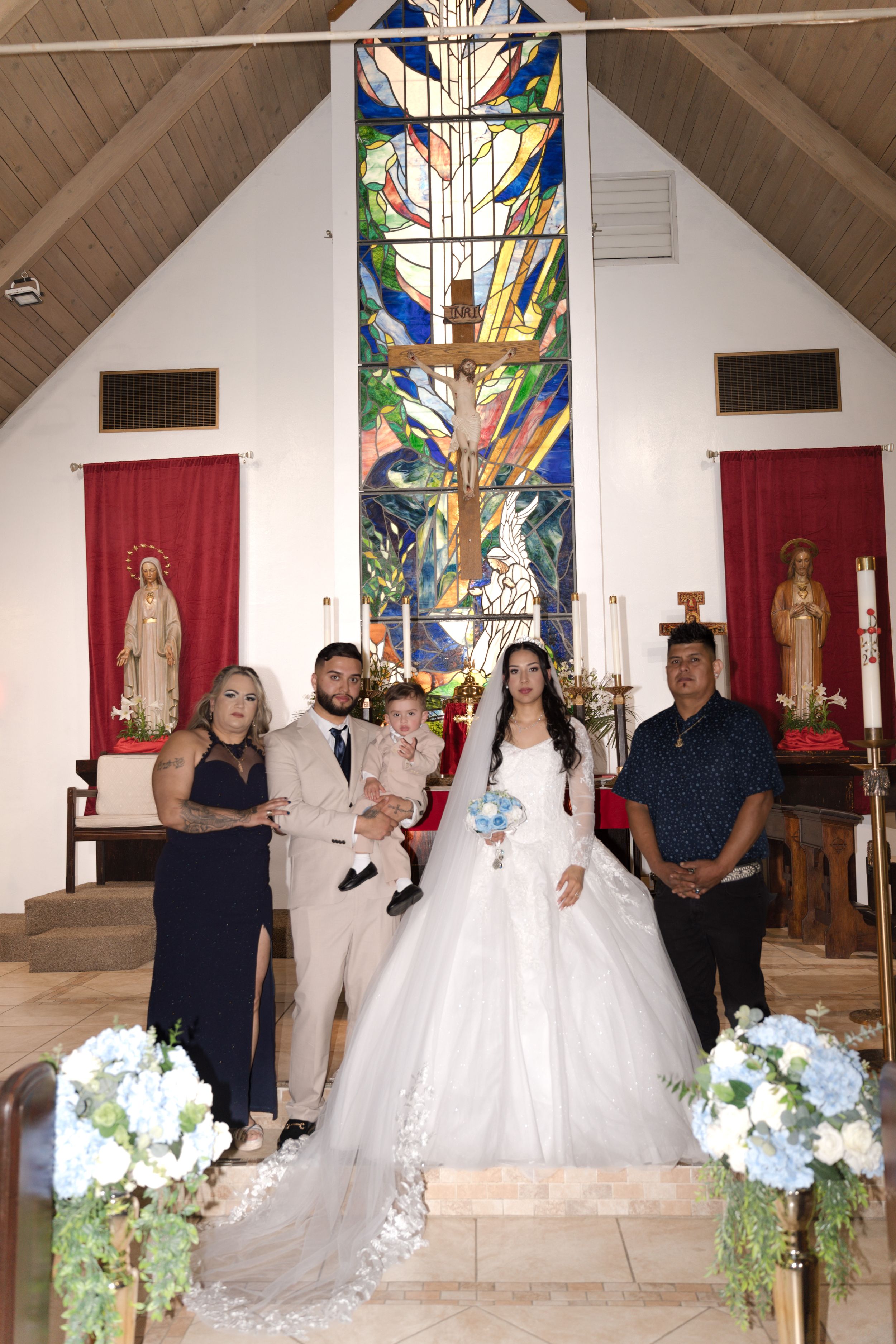 Wedding couple poses with family in a church featuring a colorful stained glass window and floral arrangements.