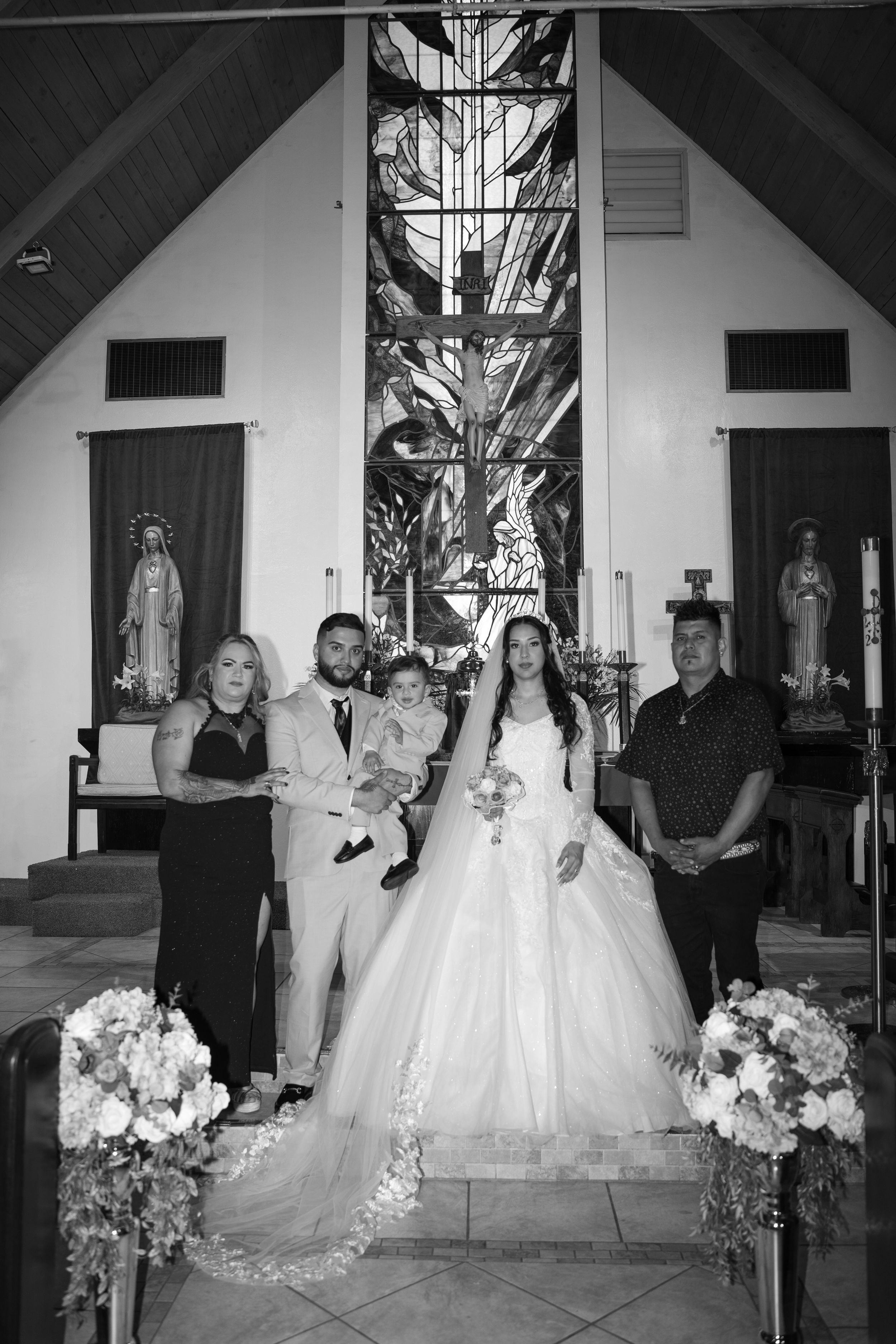 Black and white wedding photo of bride and groom with family in a Catholic church, featuring stained glass and floral arrangements.