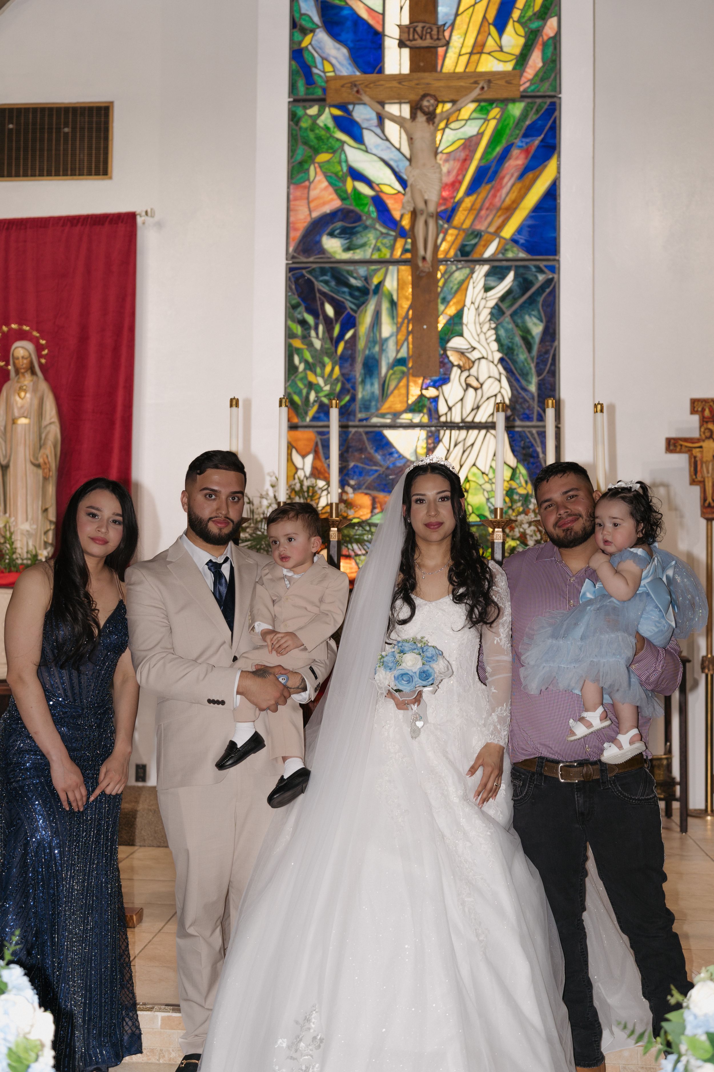 Wedding party poses before colorful stained glass cross in church, bride in white gown holds bouquet, groom in tan suit holds toddler.