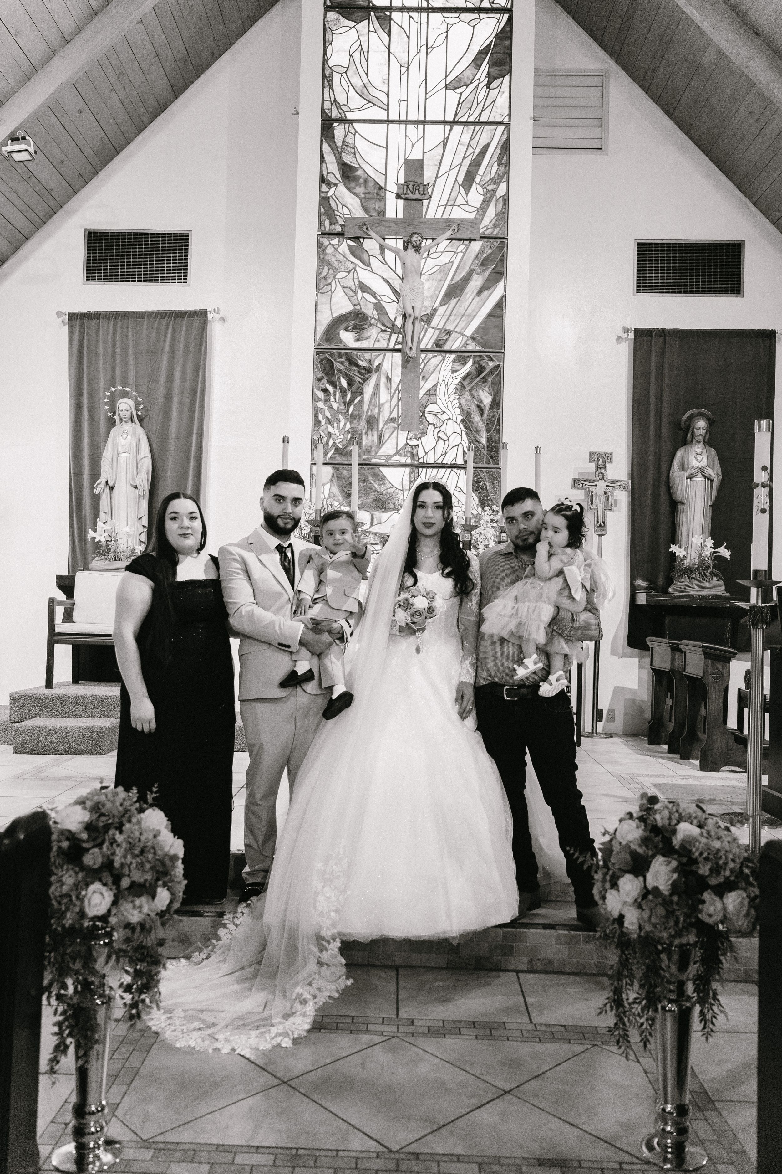 Black and white wedding photo of bride and groom with family posing inside a decorated church altar.