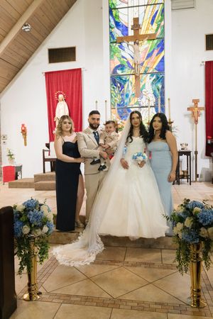Wedding party posing in a church with stained glass, bride in white gown, groom in tan suit, blue floral arrangements.