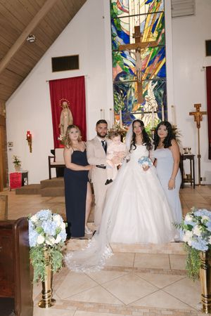 Wedding party posing at church altar with stained glass window, bride in white gown, groom in tan suit, bridesmaids in blue.