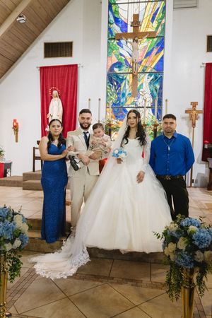 Wedding party posing at church altar with stained glass cross, bride in white gown, guests in blue attire holding baby.