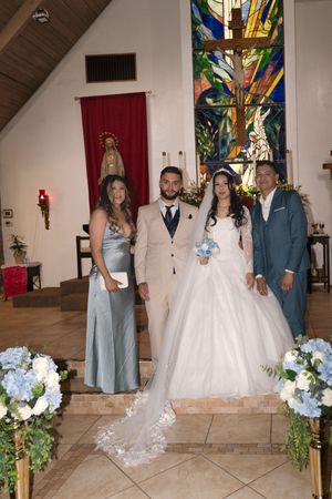 Bride and groom pose with two guests inside a church decorated with stained glass, floral arrangements, and a crucifix.