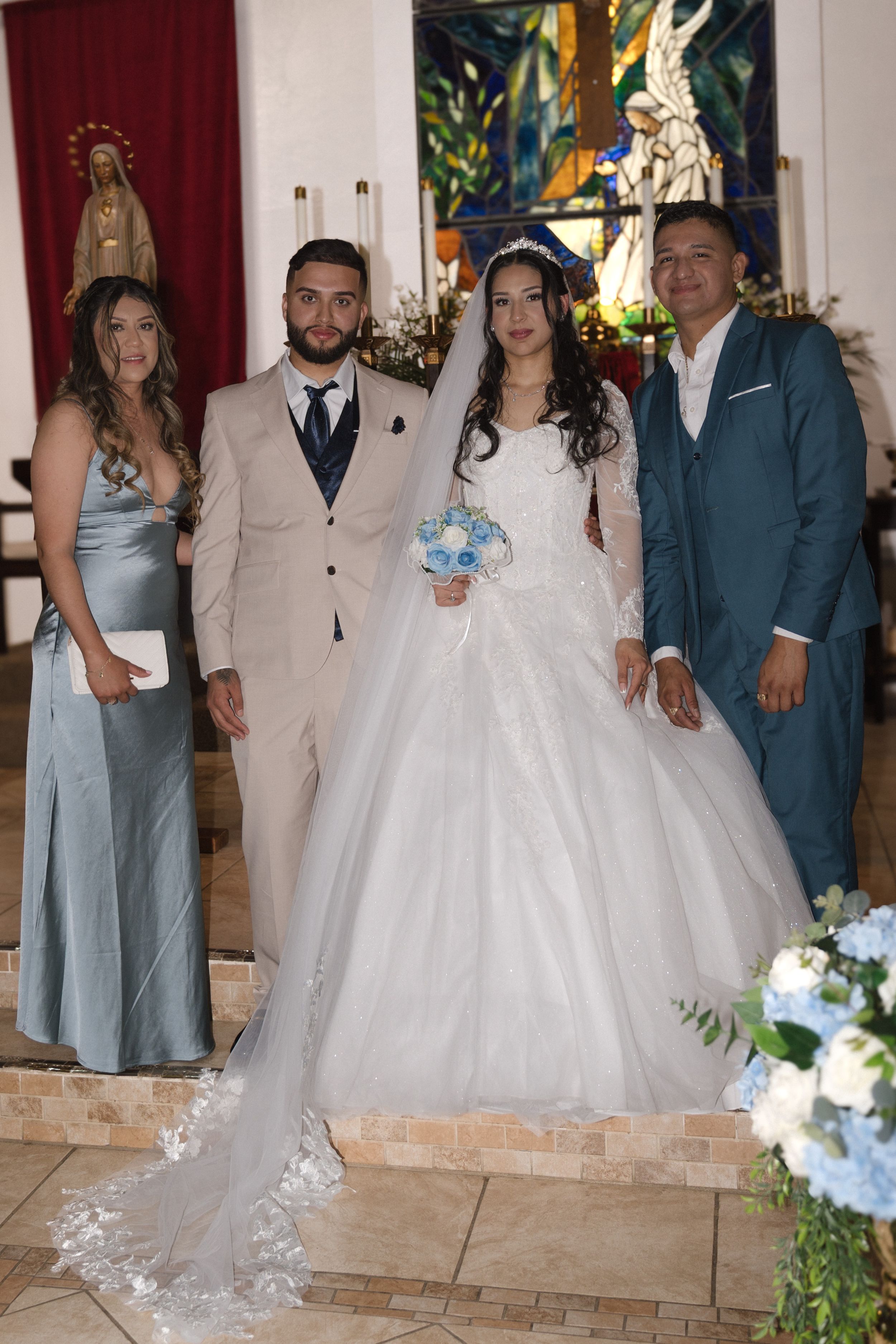Bride in white ballgown and groom in beige suit pose with guests inside a decorated church wedding ceremony.