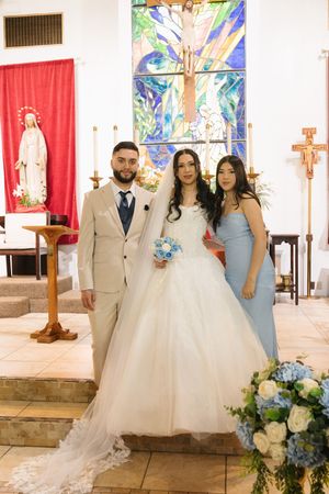 Bride and groom pose with bridesmaid at church altar with stained glass window and blue floral arrangements.