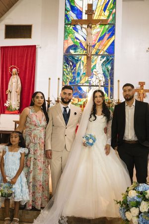 Bride and groom pose with family in front of stained glass cross at Catholic church wedding ceremony.