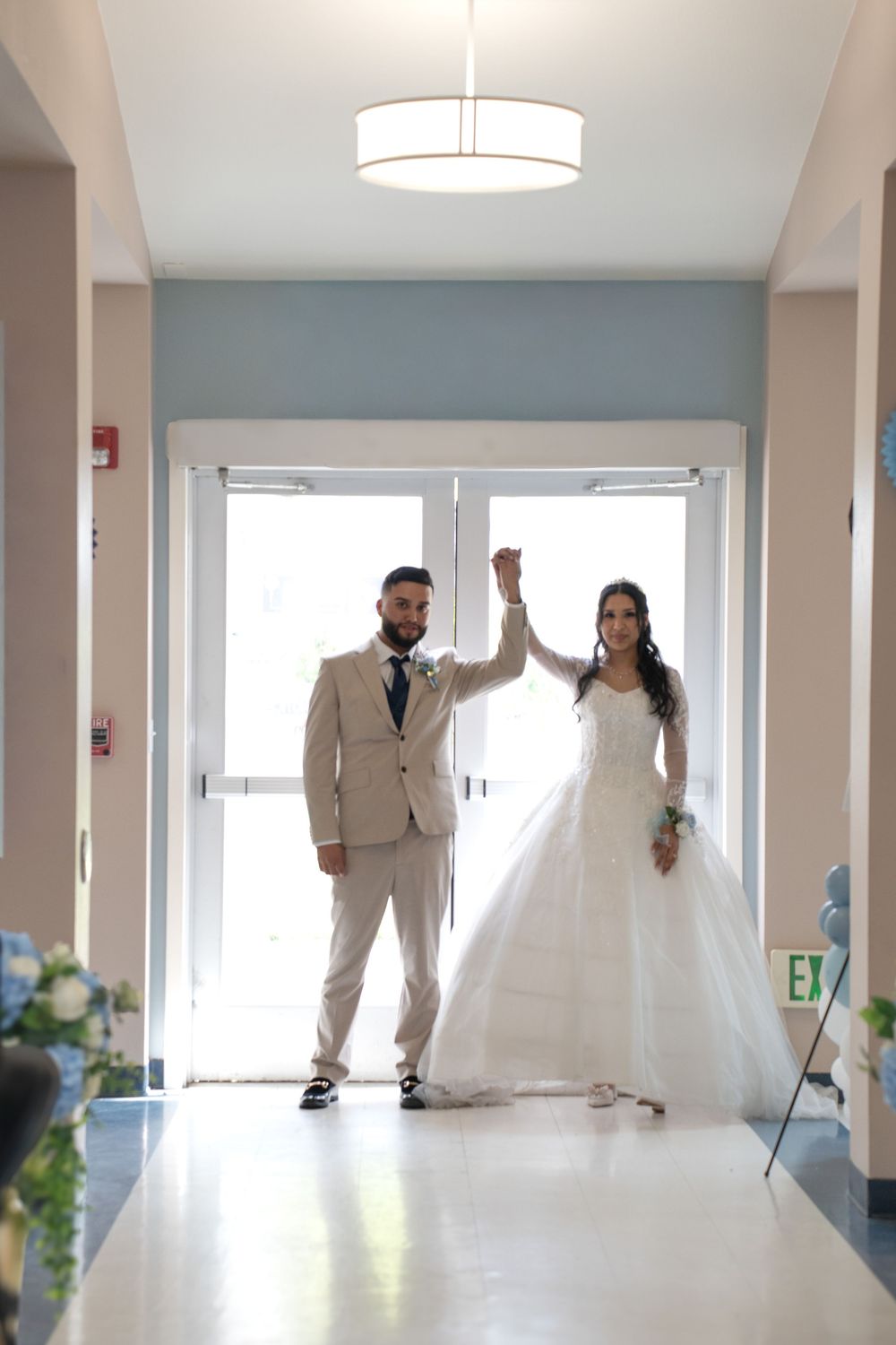 Bride and groom holding hands raised in a hallway, wearing white gown and beige suit, celebrating their wedding day.