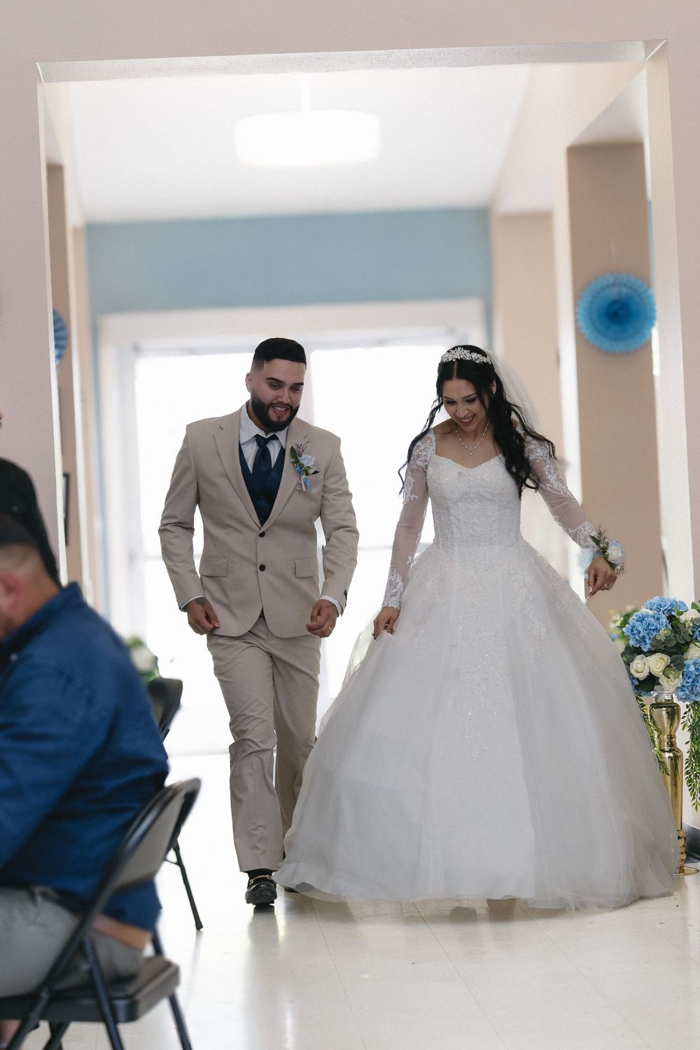 Bride in white ballgown and groom in tan suit walk together down aisle at wedding ceremony.