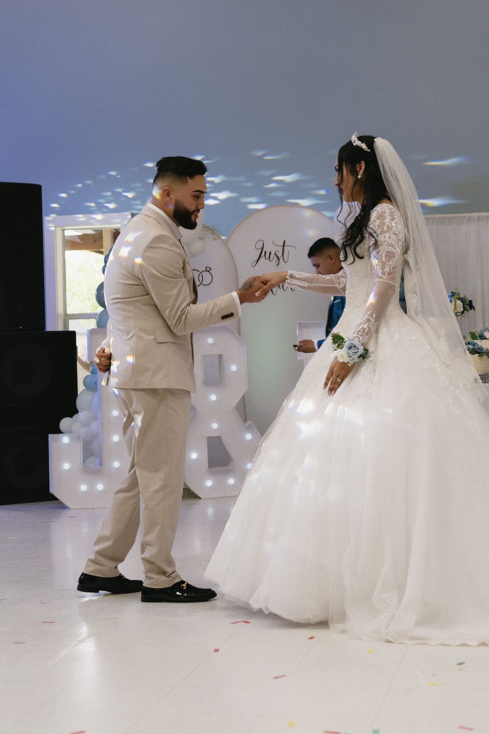 Bride and groom hold hands during wedding ceremony on white dance floor with Just Married backdrop and illuminated letters.