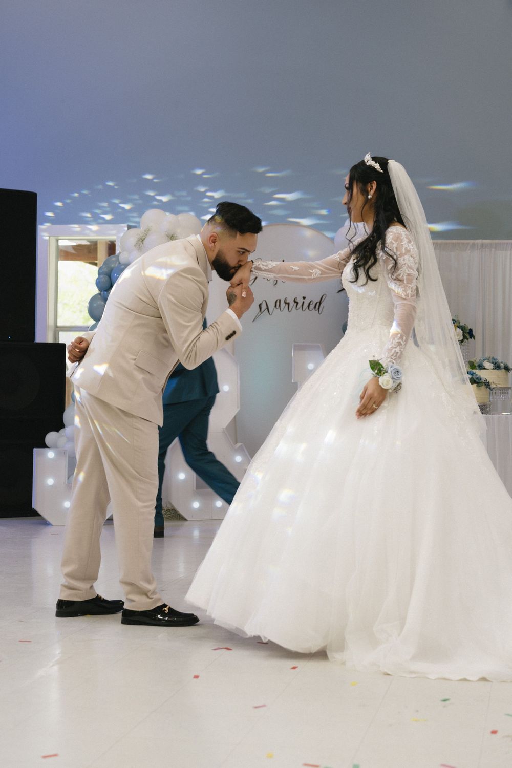 Groom in beige suit kissing bride's hand during first dance at elegant wedding reception.