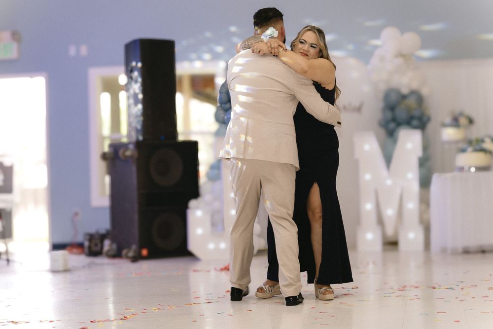 Couple sharing an emotional dance floor hug at a formal event with decorative lights and letter M backdrop.