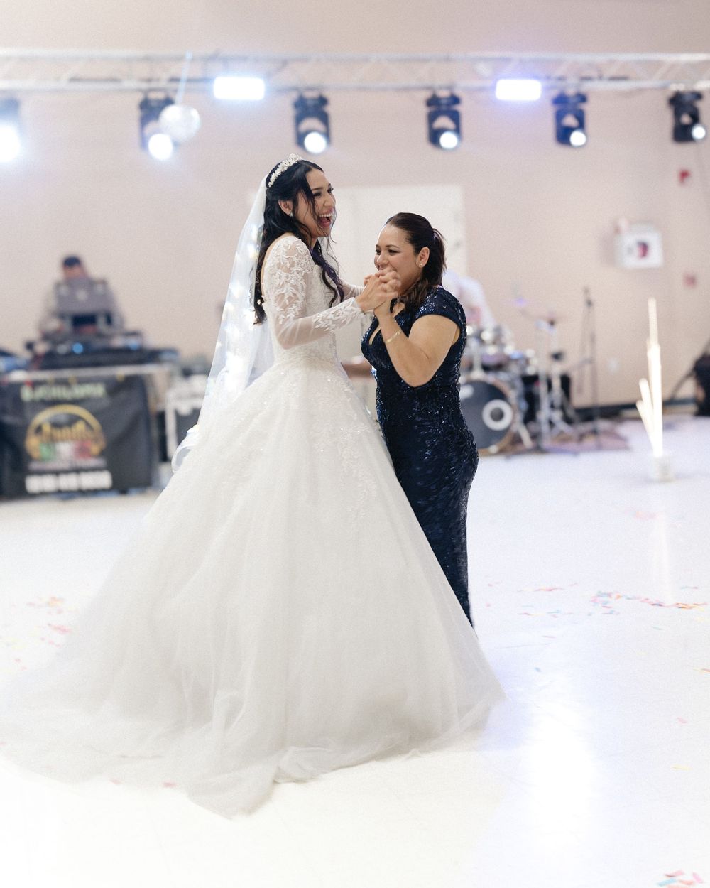 Bride in white ball gown shares emotional dance with woman in black sequin dress at wedding reception.